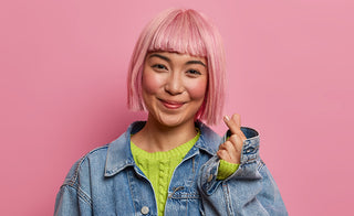 Smiling Asian girl with pink dyed bob hair makes Korean sign of love, fingers shaped into heart symbol, wears oversized denim jacket, stands against pink studio background