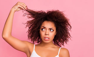 African American woman touching her natural hair with a confused look on her face as she wears a white shirt standing in front of a pink background
