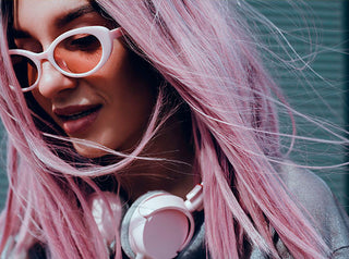 Girl with pink color treated hair and white glasses looking down with headphones around her neck