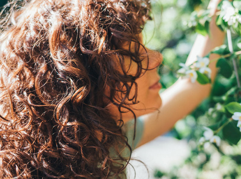 Portrait of young woman with curly frizzy hair enjoying scent of blooming tree flowers during the summer.