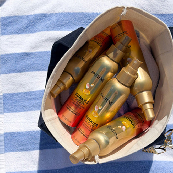 A canvas bag filled with several gold and orange bottles of Pantene Sunkissed Glow Leave-In Hair Protection Spray, a scientifically proven formula for protecting damaged hair from UV exposure, sits on a blue and white striped towel on a sandy surface.