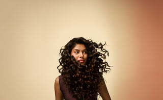 Tan-skinned woman with thick full curly brown hair looks to the side against an amber background, showing how she uses Pantene products to reduce hair loss from the root and from breakage.
