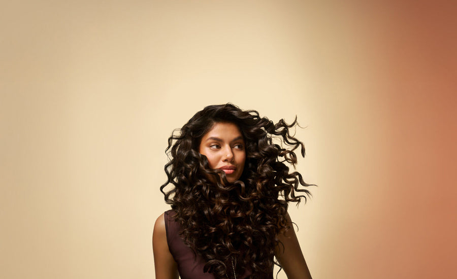Tan-skinned woman with thick full curly brown hair looks to the side against an amber background, showing how she uses Pantene products to reduce hair loss from the root and from breakage.