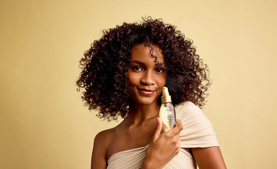 Tan-skinned woman with coily brown hair shows the amazing result on her hair after using Pantene treatments for her damaged hair in front of a gold background.