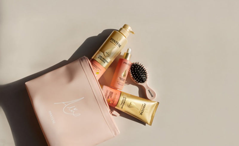 A "Alix Earle x Pantene" branded pink cosmetic bag is unzipped with “Abundant & Strong” shampoo, conditioner, serum, and a pink hairbrush inside, revealing Pantene's new innovation for hair loss treatment. 