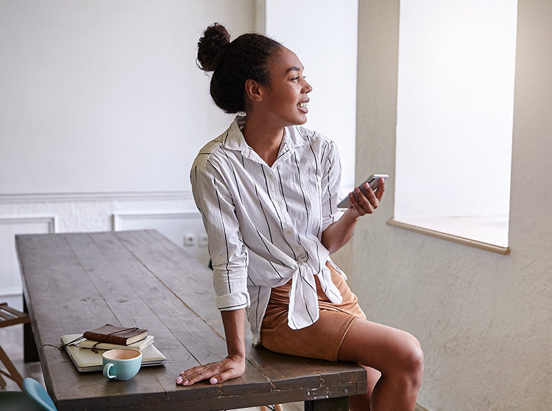 Happy dark skinned woman making break from her work, sitting on table while having pleasant talk on mobile phone, wearing white striped shirt 